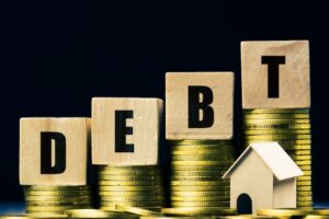Wooden blocks spelling debt upward on stacked coins with a wooden house