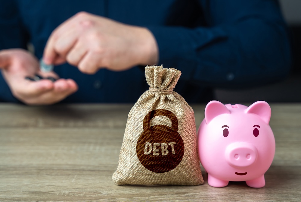 Person counting change with a bag that says debt and a piggy bank next to it