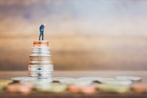 Miniature person standing on top of a stack of coins looking down on money